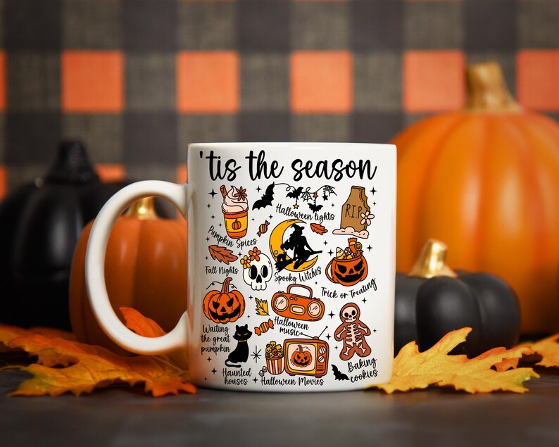 Pu&ograve; includere: Tazza in ceramica bianca con la scritta "'tis the season" e illustrazioni a tema Halloween. La tazza presenta immagini di zucche, pipistrelli, una strega e altri elementi spettrali. La tazza &egrave; circondata da zucche e foglie autunnali.