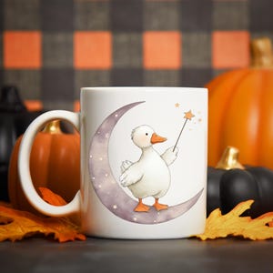 May include: White ceramic mug featuring a whimsical illustration of a white duck holding a star wand while perched on a crescent moon. The design includes stars and is set against a backdrop of pumpkins and fall leaves.