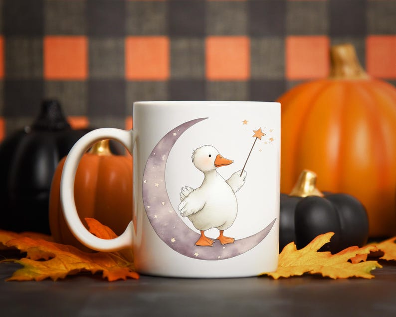 May include: White ceramic mug featuring a whimsical illustration of a white duck holding a star wand while perched on a crescent moon. The design includes stars and is set against a backdrop of pumpkins and fall leaves.