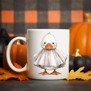 May include: White ceramic mug featuring a cartoon duck dressed as a ghost, with a light gray sheet. The duck has an orange beak and feet, with a Halloween theme. Pumpkins and fall leaves are in the background.