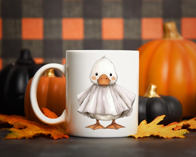 May include: White ceramic mug featuring a cartoon duck dressed as a ghost, with a light gray sheet. The duck has an orange beak and feet, with a Halloween theme. Pumpkins and fall leaves are in the background.