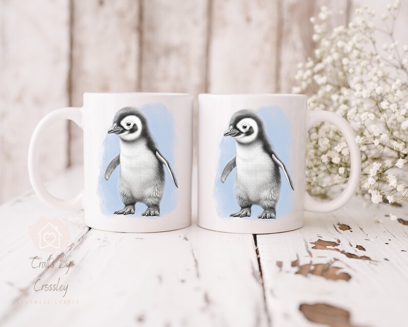 May include: Two white ceramic mugs with a blue watercolour background and a black and white illustration of a penguin chick on each mug.