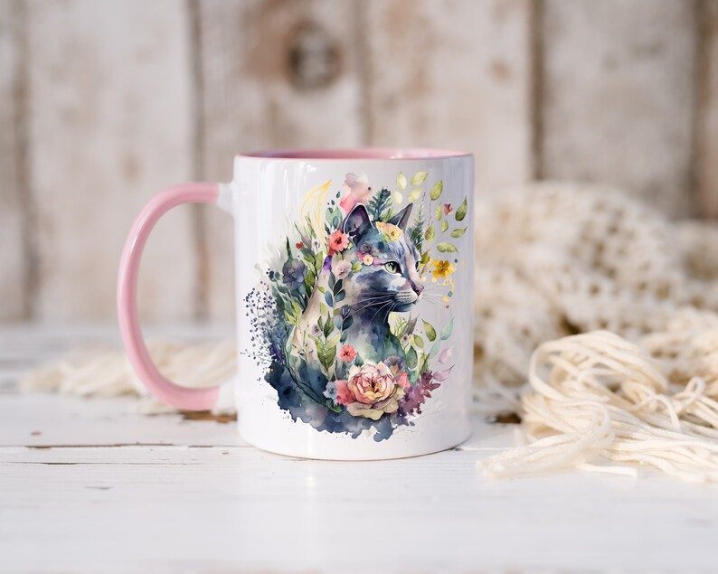 May include: White ceramic mug with a pink handle and a watercolor design of a cat surrounded by flowers. The cat is blue and gray with green eyes.