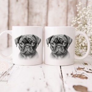 May include: Two white ceramic mugs with black and white illustrations of a pug puppy on each side. The mugs are on a white wooden surface.