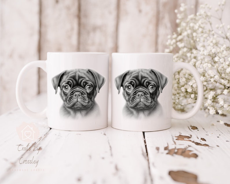 May include: Two white ceramic mugs with black and white illustrations of a pug puppy on each side. The mugs are on a white wooden surface.
