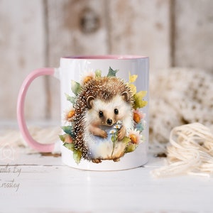 May include: A white ceramic mug with a pink rim and handle. The mug features a watercolor illustration of a hedgehog holding a bouquet of white flowers. The hedgehog is surrounded by green leaves and flowers.