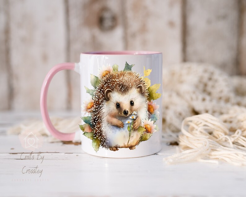 May include: A white ceramic mug with a pink rim and handle. The mug features a watercolor illustration of a hedgehog holding a bouquet of white flowers. The hedgehog is surrounded by green leaves and flowers.