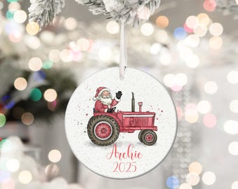 May include: A round Christmas ornament featuring Santa Claus driving a red tractor. The ornament has the name "Archie" and the year "2025" printed in a script font. The ornament is hanging from a white ribbon.