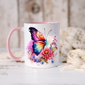 K&ouml;nnte beinhalten: Eine wei&szlig;e Keramiktasse mit rosafarbenem Rand und Henkel zeigt einen bunten Schmetterling mit einem galaktischen Muster auf seinen Fl&uuml;geln. Der Schmetterling ist von Aquarellblumen in Rosa-, Blau- und Violettt&ouml;nen umgeben.