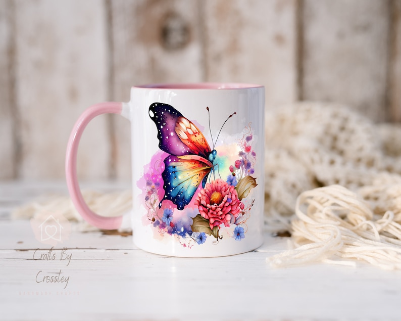 K&ouml;nnte beinhalten: Eine wei&szlig;e Keramiktasse mit rosafarbenem Rand und Henkel zeigt einen bunten Schmetterling mit einem galaktischen Muster auf seinen Fl&uuml;geln. Der Schmetterling ist von Aquarellblumen in Rosa-, Blau- und Violettt&ouml;nen umgeben.