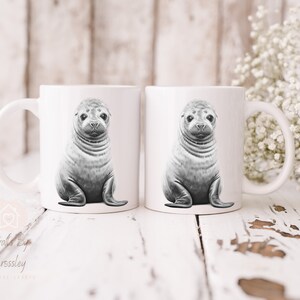 K&ouml;nnte beinhalten: Zwei wei&szlig;e Keramikbecher mit einer Schwarzwei&szlig;illustration eines Seehunde-Babys auf jedem Becher. Die Becher stehen auf einer wei&szlig;en Holzoberfl&auml;che.