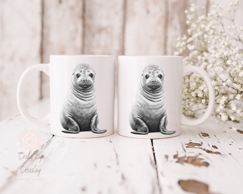 K&ouml;nnte beinhalten: Zwei wei&szlig;e Keramikbecher mit einer Schwarzwei&szlig;illustration eines Seehunde-Babys auf jedem Becher. Die Becher stehen auf einer wei&szlig;en Holzoberfl&auml;che.