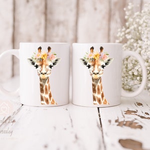 May include: Two white ceramic mugs with a giraffe wearing a flower crown printed on them. The giraffe is brown and white with a light brown background.