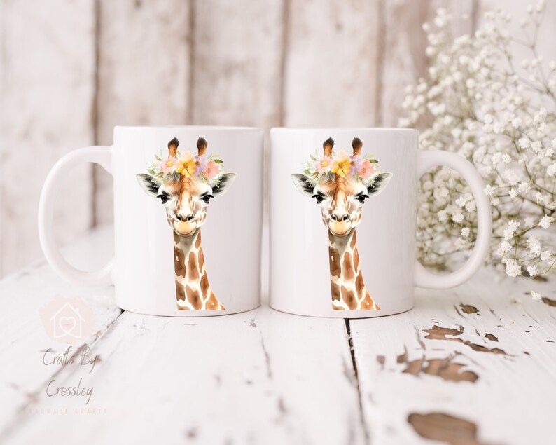 May include: Two white ceramic mugs with a giraffe wearing a flower crown printed on them. The giraffe is brown and white with a light brown background.