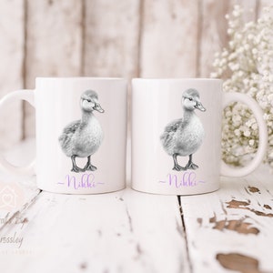 May include: Two white ceramic mugs featuring a black and white illustration of a duckling on each. The mugs have the name "Nikki" written in purple at the bottom.