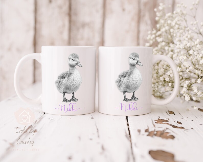 May include: Two white ceramic mugs featuring a black and white illustration of a duckling on each. The mugs have the name "Nikki" written in purple at the bottom.