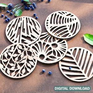 May include: Five round wooden coasters with intricate laser cut designs. The coasters feature various patterns including geometric shapes, leaves, and swirls. The coasters are arranged on a brown surface.
