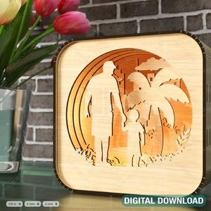 Op de afbeelding: Een nachtlampje van lasergesneden hout met een silhouet van een vader en kind dat hand in hand over een strand met palmbomen loopt. Het ontwerp wordt van achteren verlicht, waardoor een warm gloed ontstaat.