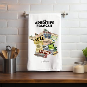 May include: White kitchen towel with the text "LES APÉRITIFS FRANÇAIS" above a map of France filled with vintage liquor labels. The towel is hanging on a metal bar against a white brick wall.