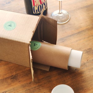 May include: A brown cardboard box with a white label that says "Adventure awaits" and a green triangle logo. The box is open and a brown tube with a white end is partially inside. A clear glass wine bottle with a red and white label that says "Easy Going" is sitting on top of the box. A clear wine glass is sitting on the table next to the box.