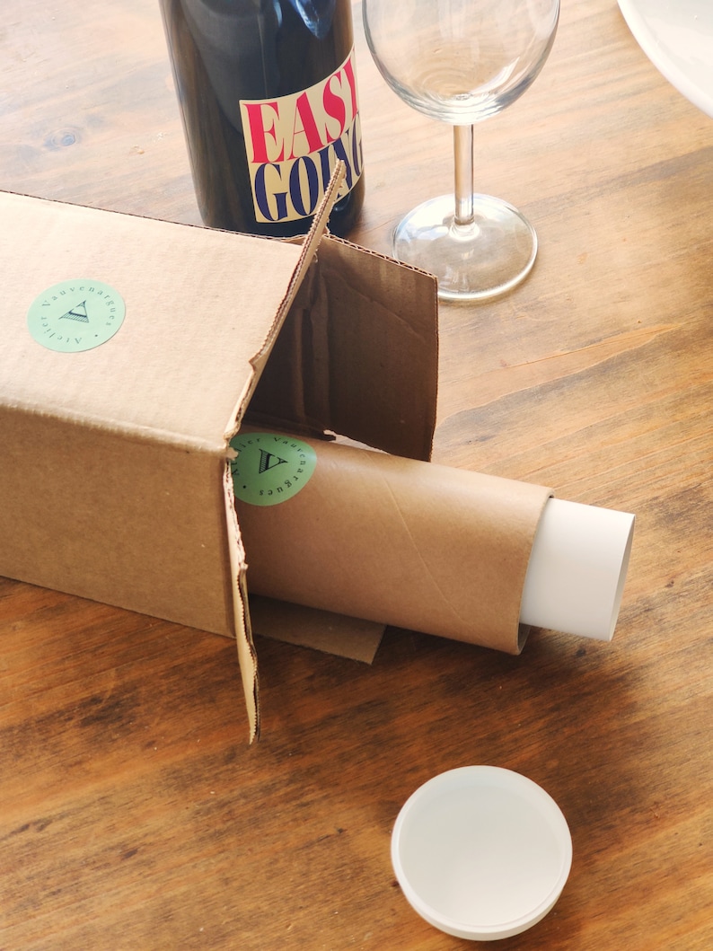 May include: A brown cardboard box with a white label that says "Adventure awaits" and a green triangle logo. The box is open and a brown tube with a white end is partially inside. A clear glass wine bottle with a red and white label that says "Easy Going" is sitting on top of the box. A clear wine glass is sitting on the table next to the box.
