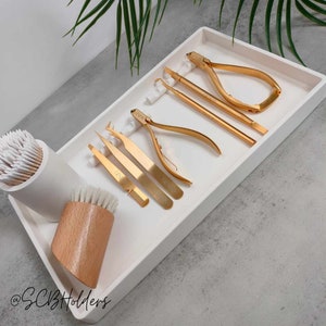 Pode incluir: Uma bandeja branca com um conjunto de ferramentas douradas para unhas, incluindo pin&ccedil;as, cortadores de unhas e um empurrador de cut&iacute;culas. H&aacute; tamb&eacute;m uma escova de madeira e um recipiente branco com cotonetes.