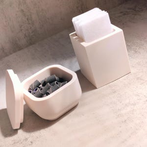 May include: Two white ceramic containers, one with a lid, the other open. The container with the lid is filled with small black and purple nail drill bits. The open container is empty.