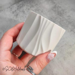 May include: A white ceramic planter with a wavy, textured design. The planter is being held in a hand with a ring that says "I am enough".