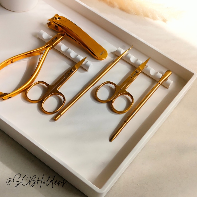 Pode incluir: Uma bandeja branca com cinco ferramentas de manicure douradas. As ferramentas incluem um cortador de unhas, pin&ccedil;as para cut&iacute;culas, dois pares de pin&ccedil;as e um empurrador de cut&iacute;culas. A bandeja tem um inserto branco com cristas para manter as ferramentas no lugar.