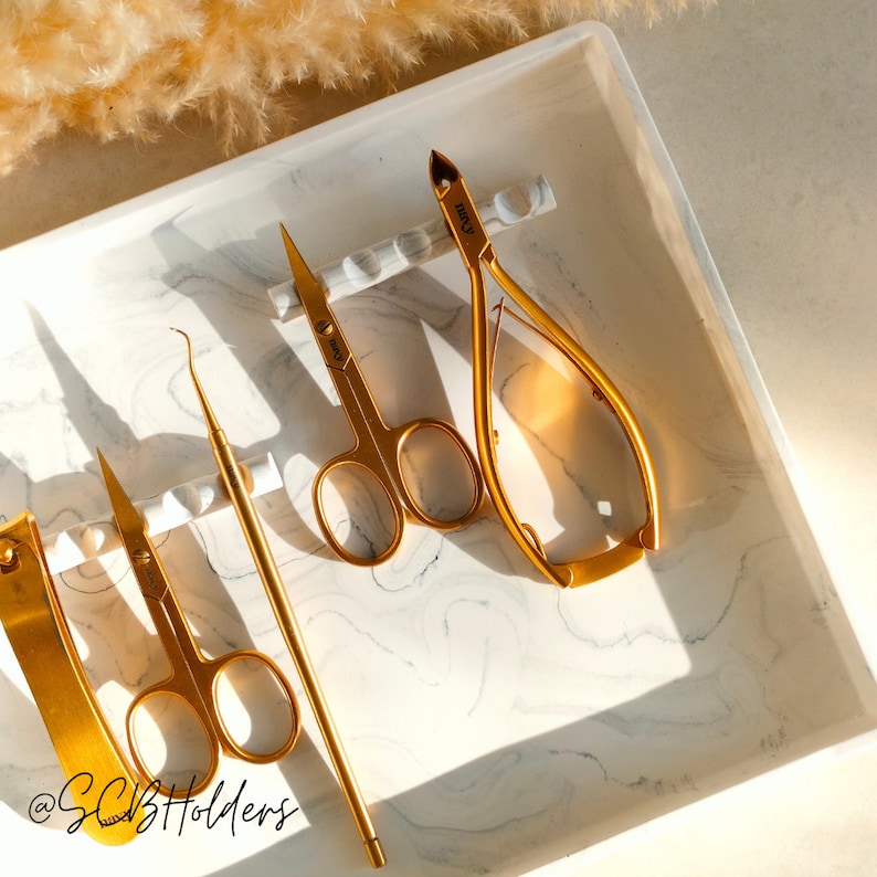 Pode incluir: Uma bandeja de m&aacute;rmore branco com quatro ferramentas de manicure douradas. As ferramentas incluem um empurrador de cut&iacute;culas, um cortador de unhas, uma pin&ccedil;a e uma lima de unhas. A bandeja tem um padr&atilde;o de m&aacute;rmore branco e as ferramentas est&atilde;o dispostas em fila.