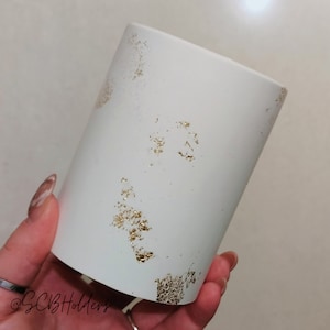 May include: White ceramic cylinder vase with gold flecks scattered across the surface. The vase is being held in a hand.