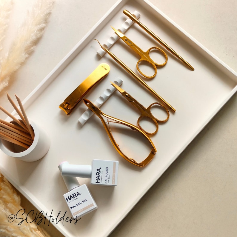 Pode incluir: Uma bandeja branca com ferramentas de manicure douradas, incluindo um cortador de unhas, um empurrador de cut&iacute;culas, pin&ccedil;as para unhas e pin&ccedil;as. Tamb&eacute;m h&aacute; duas garrafas de esmalte de unhas na bandeja, uma etiquetada como "HARA BUILDER GEL" e a outra como "HARA GEL POLISH".