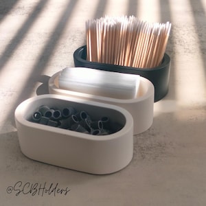 May include: Three oval containers in black and white, holding wooden sticks, white squares, and grey objects. The containers are on a light grey surface, with shadows from blinds. The text "@SCBHolders" is in the bottom left corner.