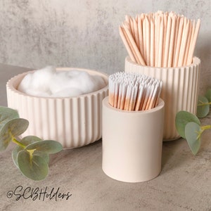 May include: Three white ceramic containers with ribbed detailing. The containers hold cotton wool balls, cotton buds, and wooden sticks. The containers are on a light grey surface with green foliage.
