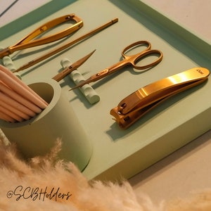 Pode incluir: Uma bandeja verde claro com um cortador de unhas dourado, uma pin&ccedil;a, um empurrador de cut&iacute;culas, uma tesoura para unhas e um suporte para limas de unhas. A bandeja est&aacute; sobre uma superf&iacute;cie branca.
