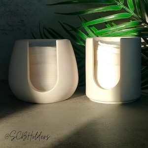 May include: Two ceramic cotton wool bud holders, one round and one oval shaped, both in a light beige colour. The holders are filled with cotton rounds. The holders are sitting on a grey surface.
