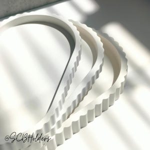 May include: Three white, round trays with a scalloped edge are stacked in a close-up shot. The top tray partially covers the others. The trays are on a white surface, with shadows. The trays are likely for serving.