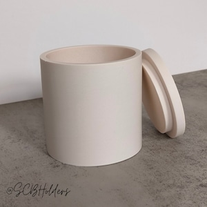 May include: A cream-coloured cylindrical container with a lid. The container is made of a smooth, matt material. The lid is slightly ajar, revealing the interior of the container.