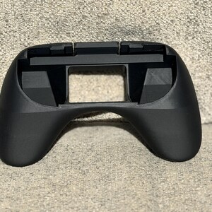 May include: A black ergonomic controller shell with a central rectangular opening and curved handles. The top features two raised sections. The device is designed for comfortable handling during extended use.
