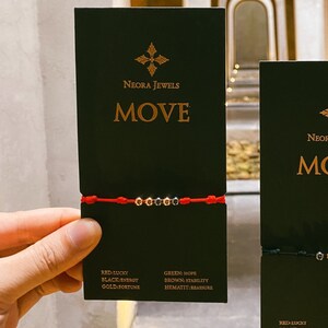 Könnte beinhalten: Zwei schwarze Karten mit dem Text "NEORA JEWELS MOVE" darauf. Jede Karte hat ein rotes und schwarzes Armband aus Schnur mit kleinen Perlen. Der Text "RED LUCKY, BLACK ENERGY, GOLD FORTUNE" und "GREEN: HOPE, BROWN STABILITY, HEMATIT: MEASURE" ist auf den Karten gedruckt.