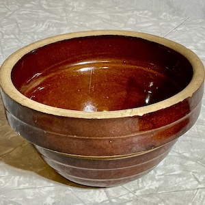 May include: A brown stoneware bowl with a light beige rim. The bowl has a glossy finish and a wide, rounded shape. The exterior features horizontal ridges. A classic piece for kitchen or home decor.