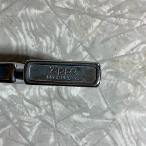 May include: Close-up of a silver Zippo lighter. The lighter's top is visible, displaying the brand name "Zippo" and the text "BRADFORD, PA." The lighter has a rectangular shape with rounded edges.