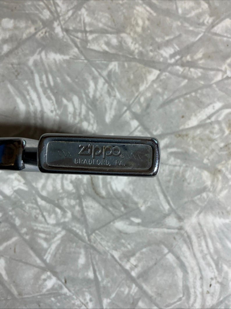 May include: Close-up of a silver Zippo lighter. The lighter's top is visible, displaying the brand name "Zippo" and the text "BRADFORD, PA." The lighter has a rectangular shape with rounded edges.
