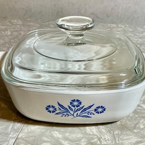 May include: White square casserole dish with a clear glass lid. The dish has a blue floral design and white handles. The lid has a clear glass knob. A vintage kitchenware item.