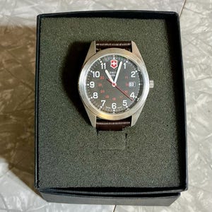 May include: A Swiss Army wristwatch with a round silver-tone stainless steel case, a black dial featuring white Arabic numerals, a red second hand, and a date window at the three o'clock position, attached to a dark brown leather strap.