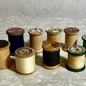 May include: A collection of vintage wooden spools of thread in various colors and sizes. Some spools are filled with black, cream, and white thread. The spools have colorful labels with text and designs. The spools are a mix of cylindrical and conical shapes.