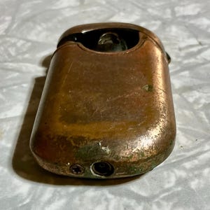 May include: A vintage copper-toned lighter with a rectangular body and rounded edges. The lighter shows signs of wear, with a tarnished finish and some green oxidation. The top features a hinged lid and a visible flint wheel.