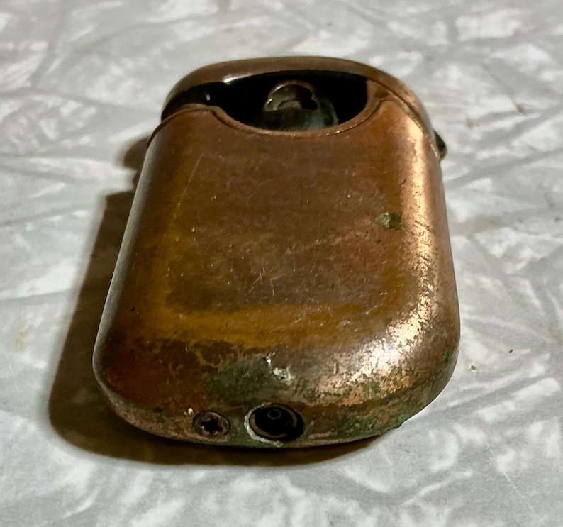 May include: A vintage copper-toned lighter with a rectangular body and rounded edges. The lighter shows signs of wear, with a tarnished finish and some green oxidation. The top features a hinged lid and a visible flint wheel.