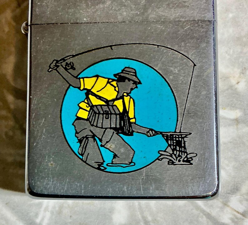 May include: A silver-colored lighter with a graphic of a fisherman in a yellow shirt and hat, set against a blue circle. The fisherman is casting a line into the water, with a fishing net visible.
