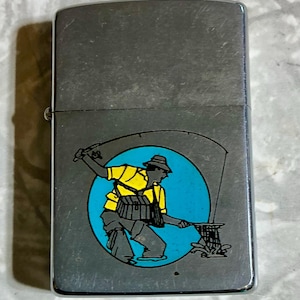 May include: A silver-toned metal lighter with a fishing-themed graphic. The graphic features a fisherman in yellow and gray attire, casting a line against a blue circle backdrop. The lighter has a rectangular shape with a hinged lid.